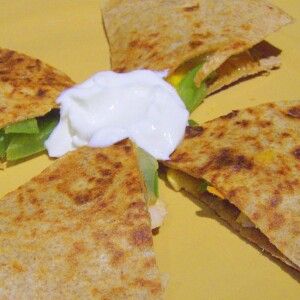 A grilled veggie quesadilla cut into quarters with sour cream in the centre. Placed on a yellow plate