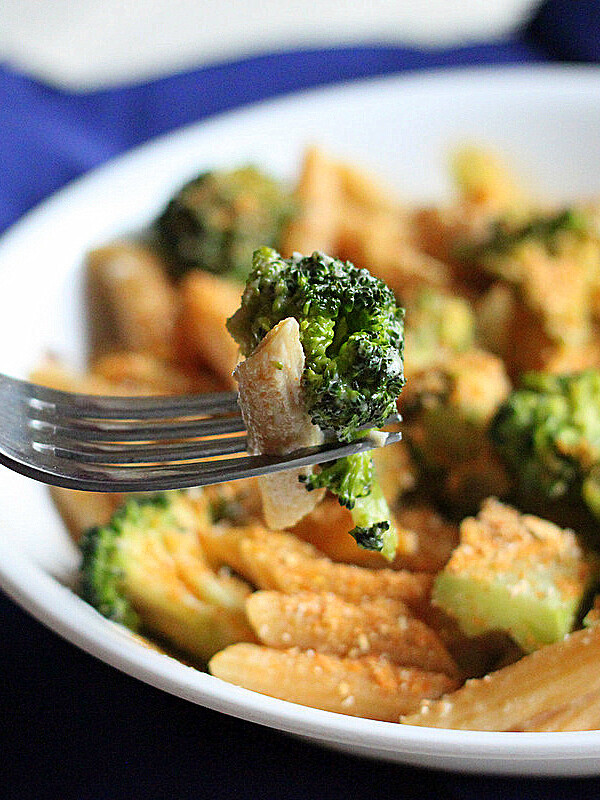 A white bowl of Penne and Broccoli in Doritos cream sauce on a blue cloth. A fork in the foreground with some penne and broccoli on it.