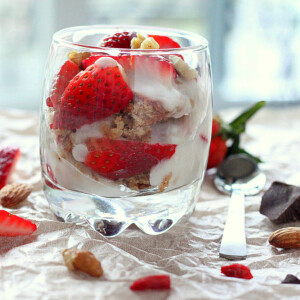 A glass of Spiced Oat Almond Gluten-free Cake, in Strawberry Nuts Goji berry parfait on a fabric background with pieces of strawberry scattered around