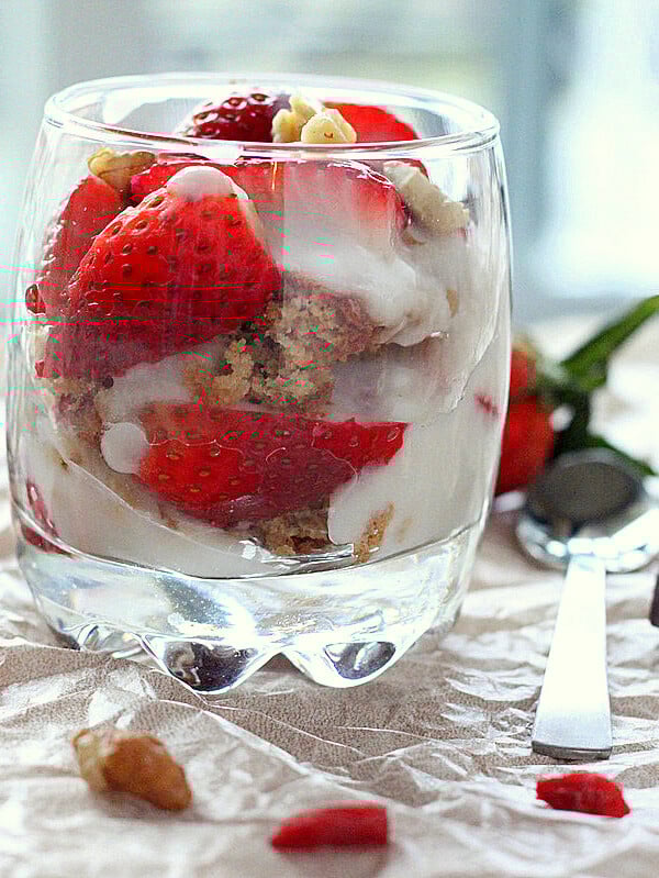 A glass of Spiced Oat Almond Gluten-free Cake, in Strawberry Nuts Goji berry parfait on a fabric background with pieces of strawberry scattered around
