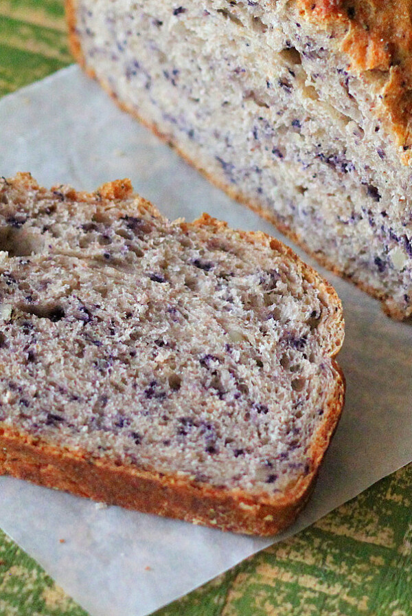 A loaf of Blueberry Almond Bread with a slice laying face up on parchemnet paper and a green background