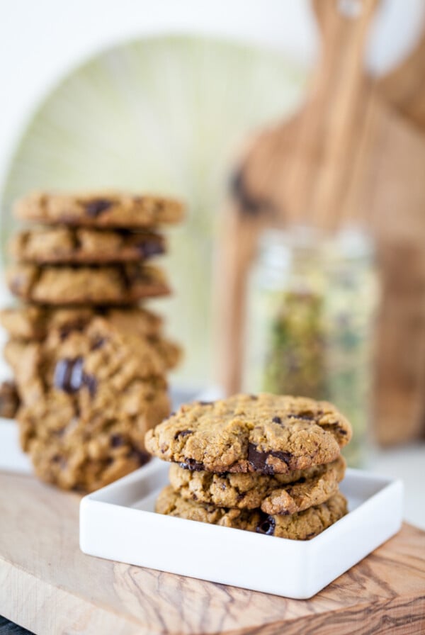 pistachio cookies on white plate with stack of vegan cookies in bakground on wooden board