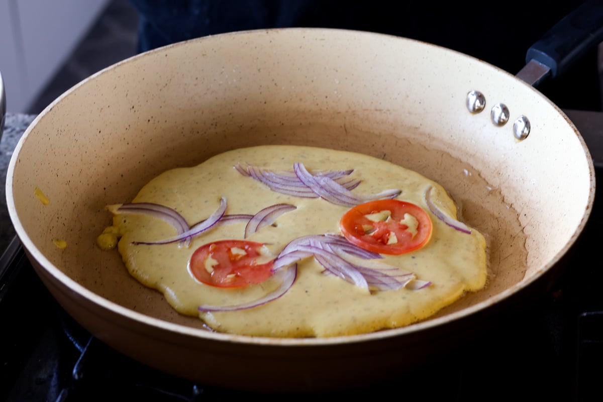 adding onion and tomato on top of the chickpea flour omelet in the pan