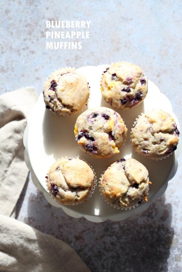 Six vegan blueberry muffins on a white plate, sitting on a pale blue worktop