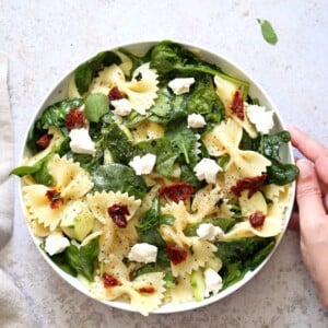 A bowl of Tuscan Pasta Salad on a light worktop. A hand holds the side of the bowl