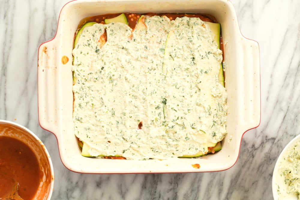 Layering the tofu ricotta on Our Vegan Gluten free Zucchini Lasagna in a cream baking dish