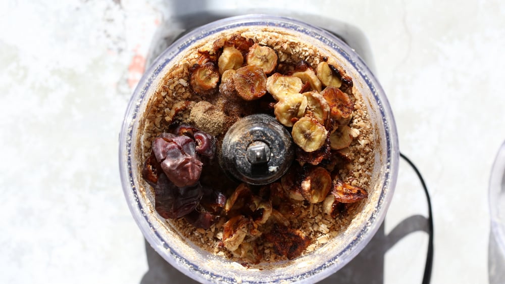 dates and bananas being added to oats and nuts in the container of a food processor
