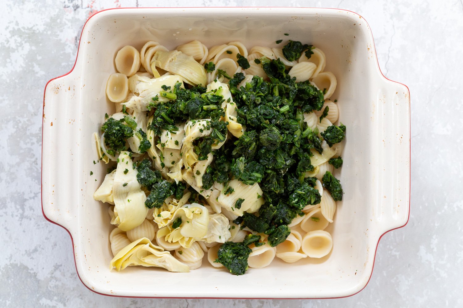 spinach pasta recipe  overhead shot of a casserole dish with cooked shell pasta, spinach, and chopped artichokes