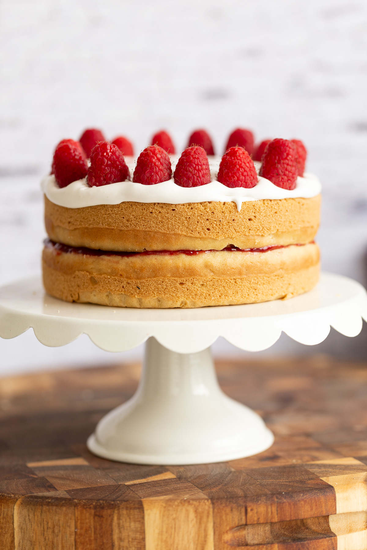 a vegan white cake topped with coconut whip and raspberries on a white cake stand