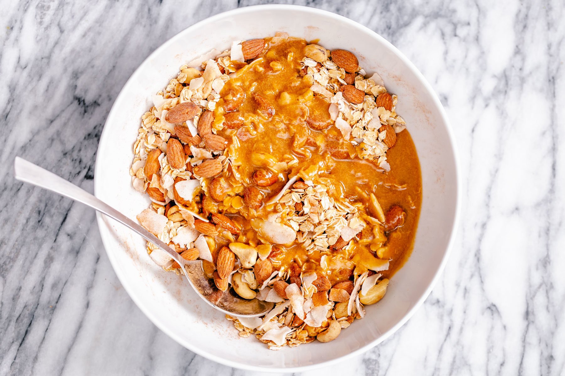 coconut granola almond butter and maple syrup being added to a small bowl with dry granola mix to make baked granola