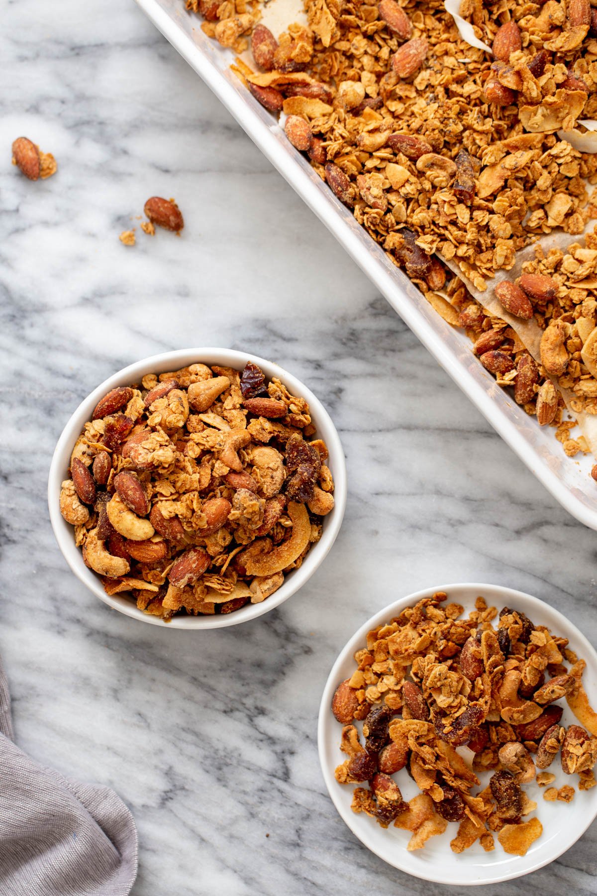 coconut granola  two small bowls of homemade baked vegan almond coconut granola next to a baking sheet with granola