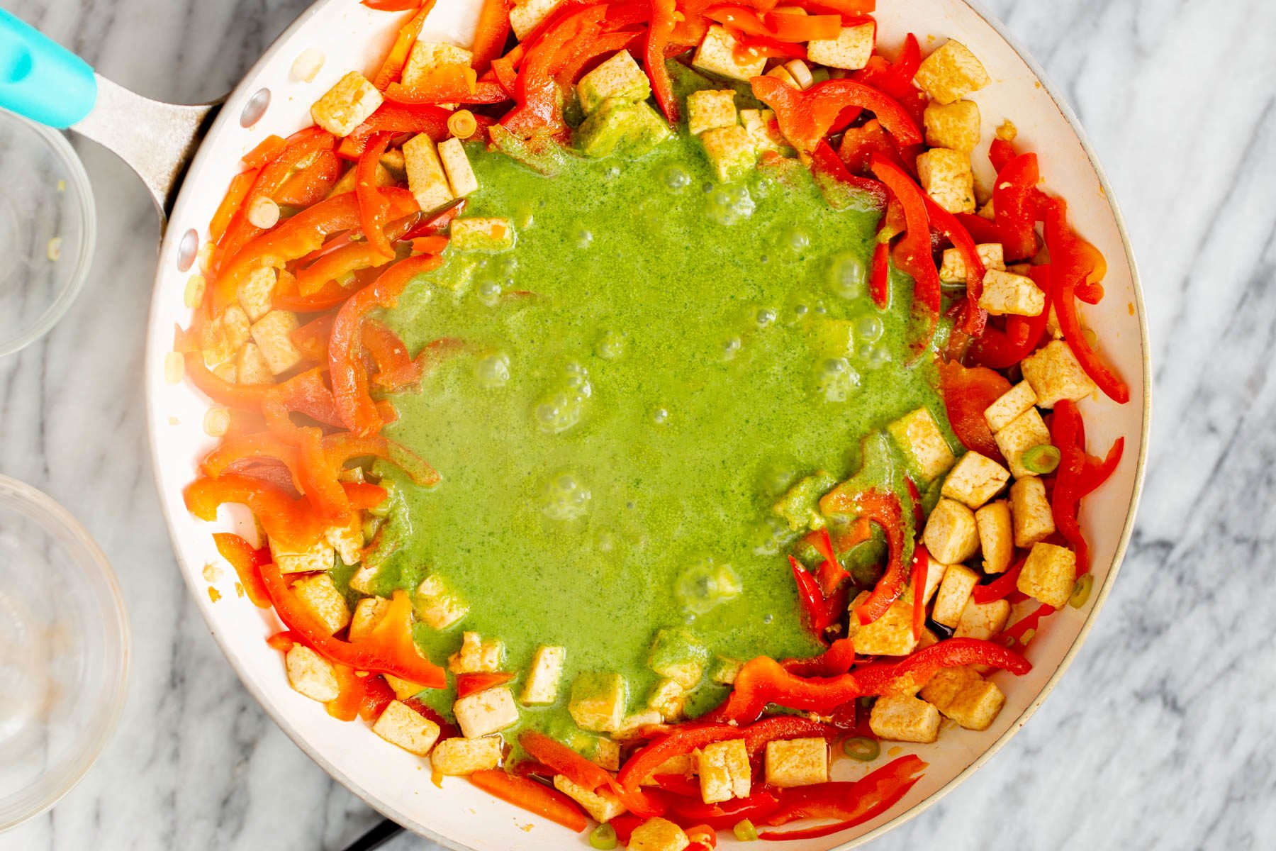 green curry sauce being poured into a frying pan with sauteed bell peppers and tofu cubes