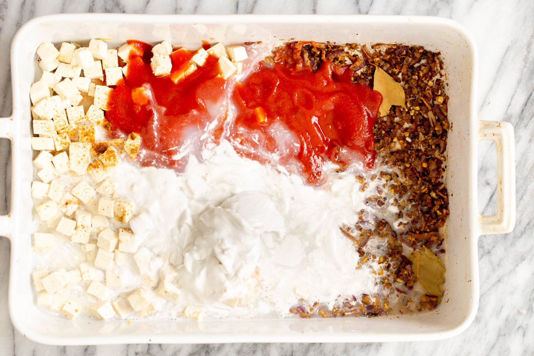 tomato puree and coconut milk being added to baked spices and tofu in casserole dish