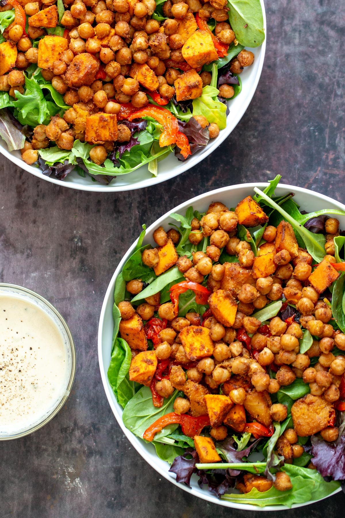 sweet potato chickpea salad two bowls with salad topped with baked chickpeas and roasted bell pepper and sweet potatoes with a small dish of tahini dressing on the side