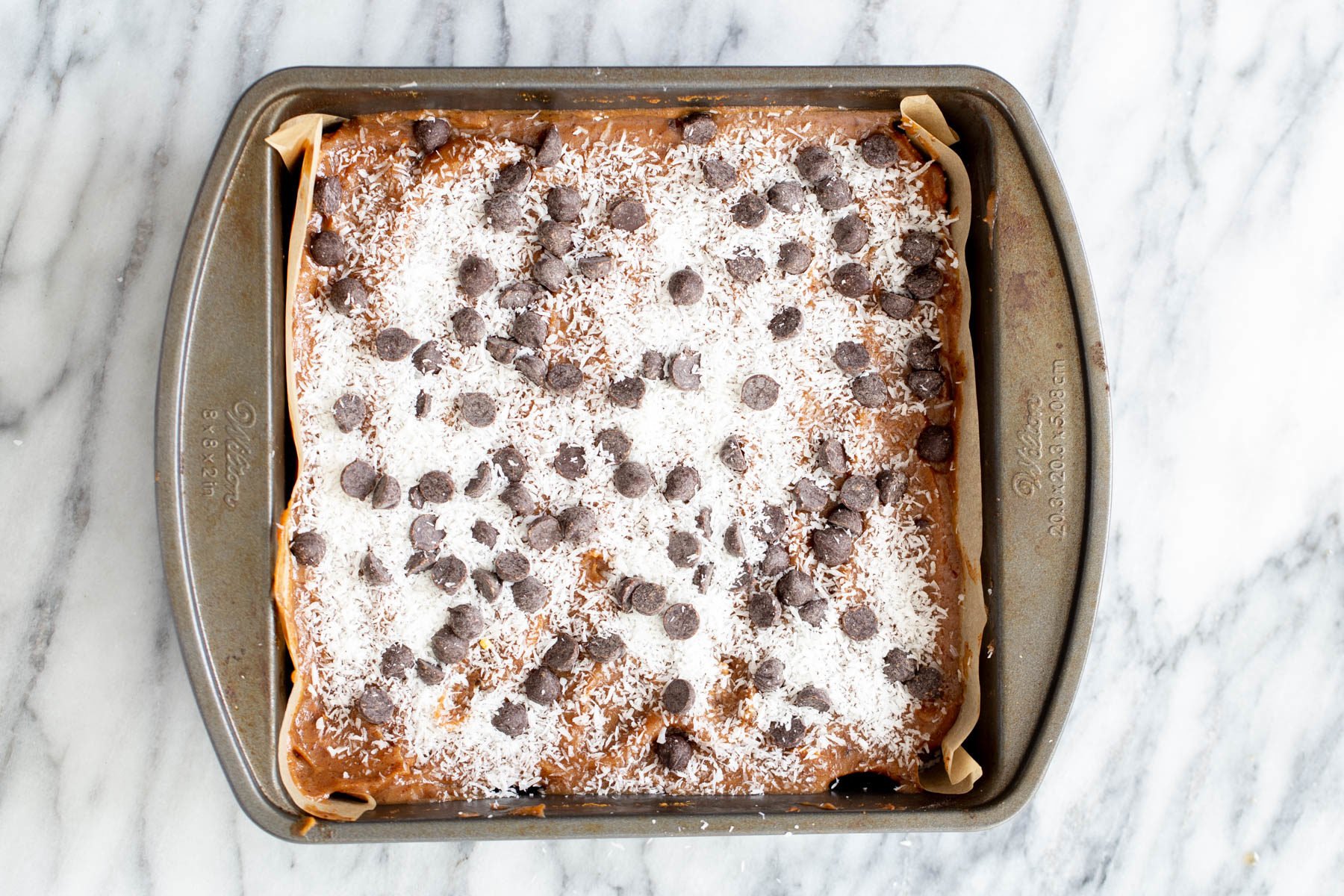 chocolate chips and shredded coconut being spread on top of oatmeal cookie base and date caramel to make caramel bars 