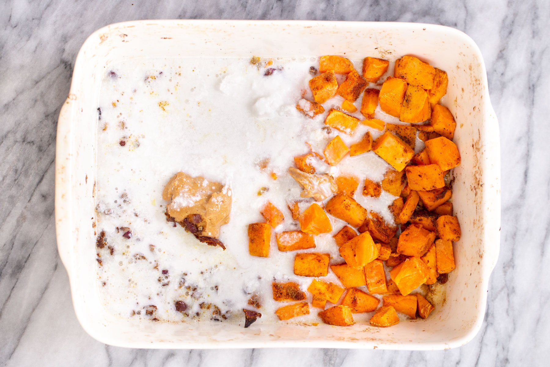 coconut milk and peanut butter being added to a casserole dish with roasted sweet potato