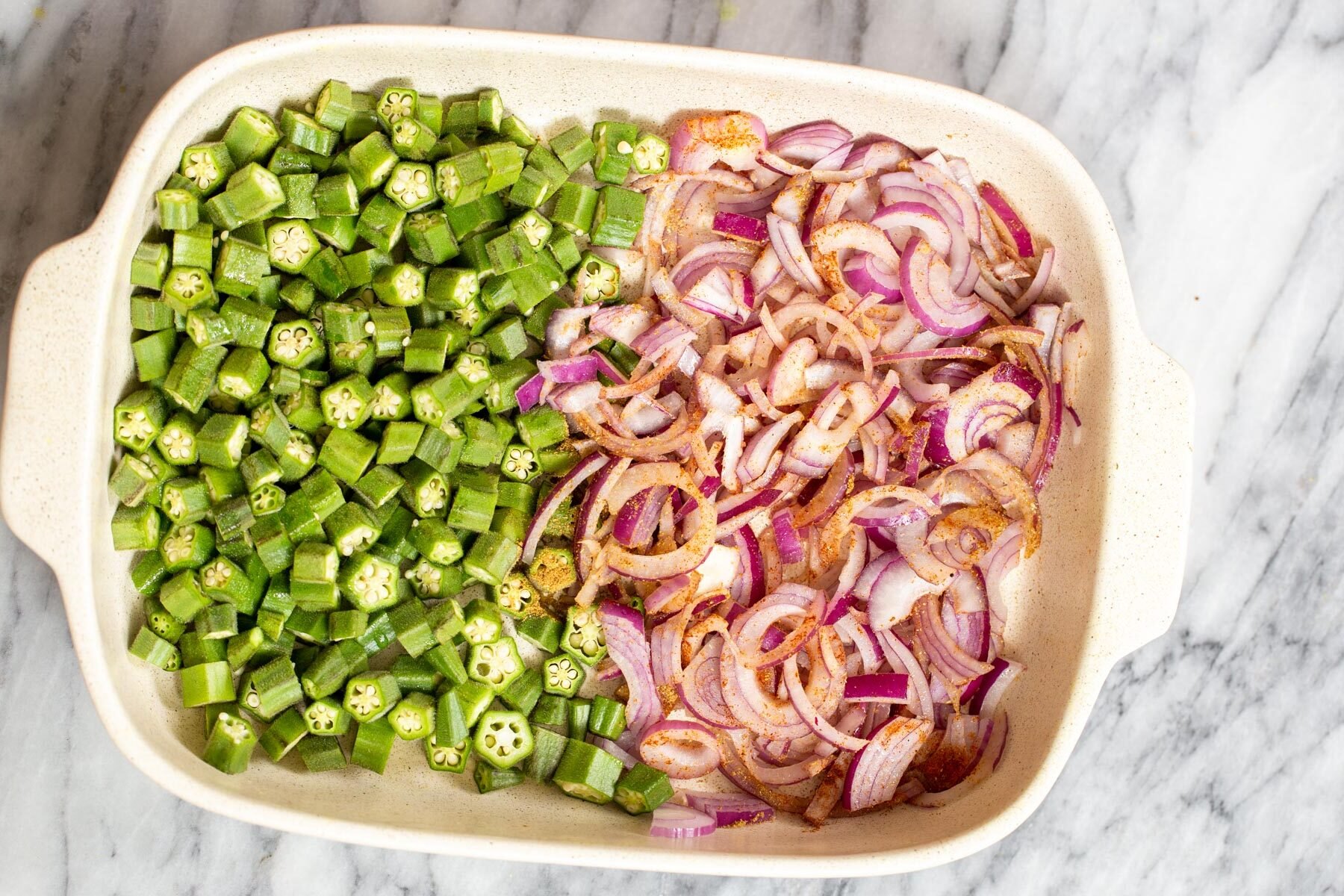 Indian spices being sprinkled over slived onion and okra in a casserole dish