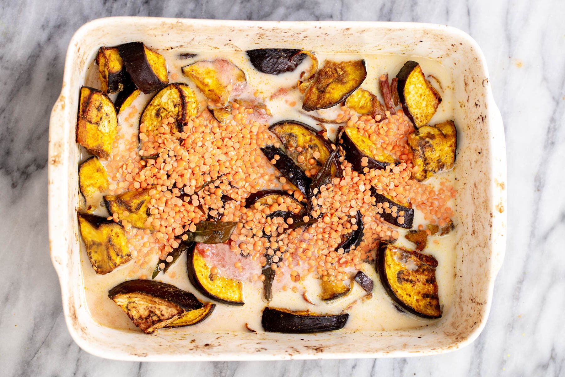 red lentils being sprinkled over eggplants in coconut milk in a white casserole dish