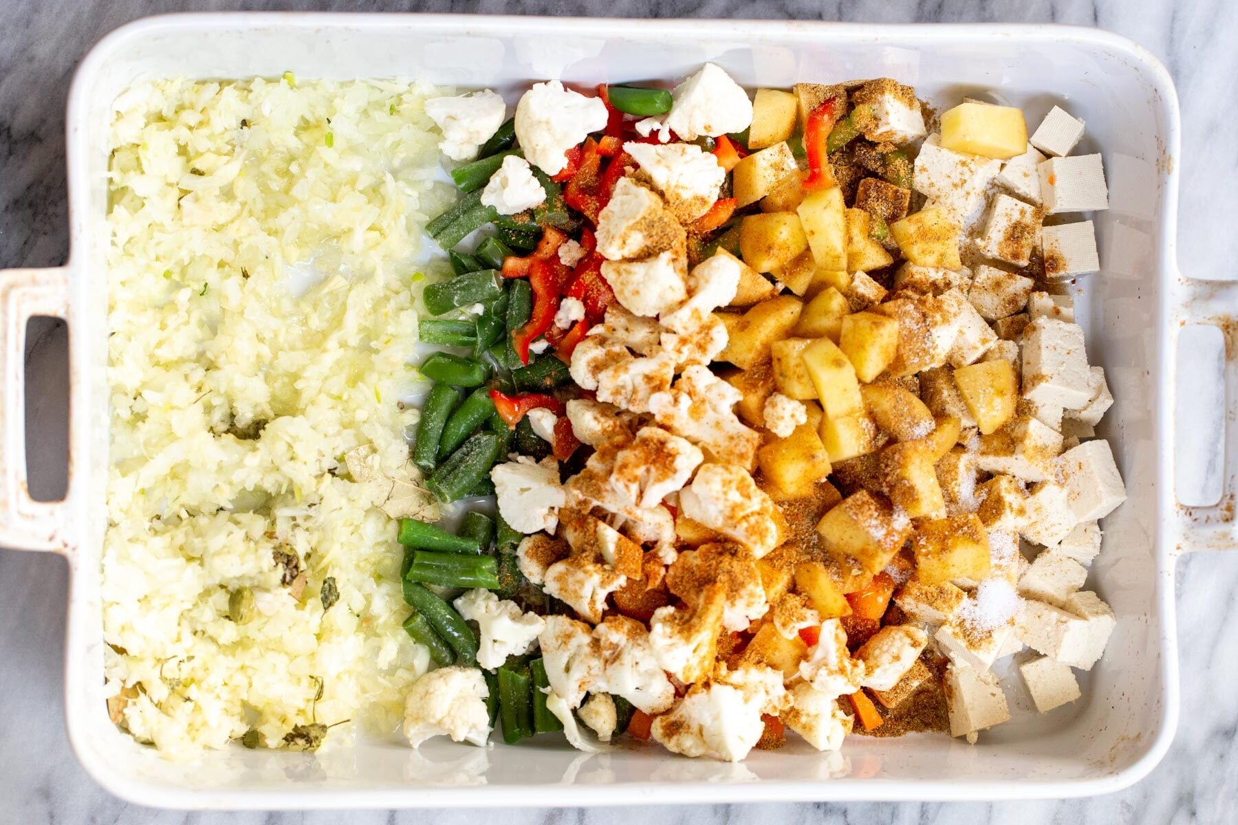 spices being sprinkled on top of veggies in a white casserole dish