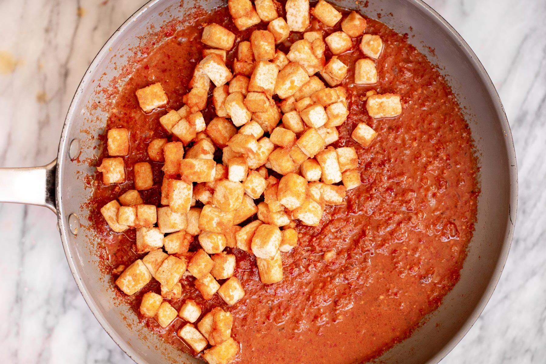 pan-fried tofu cubes being added to chili gravy in a sauteeing pan