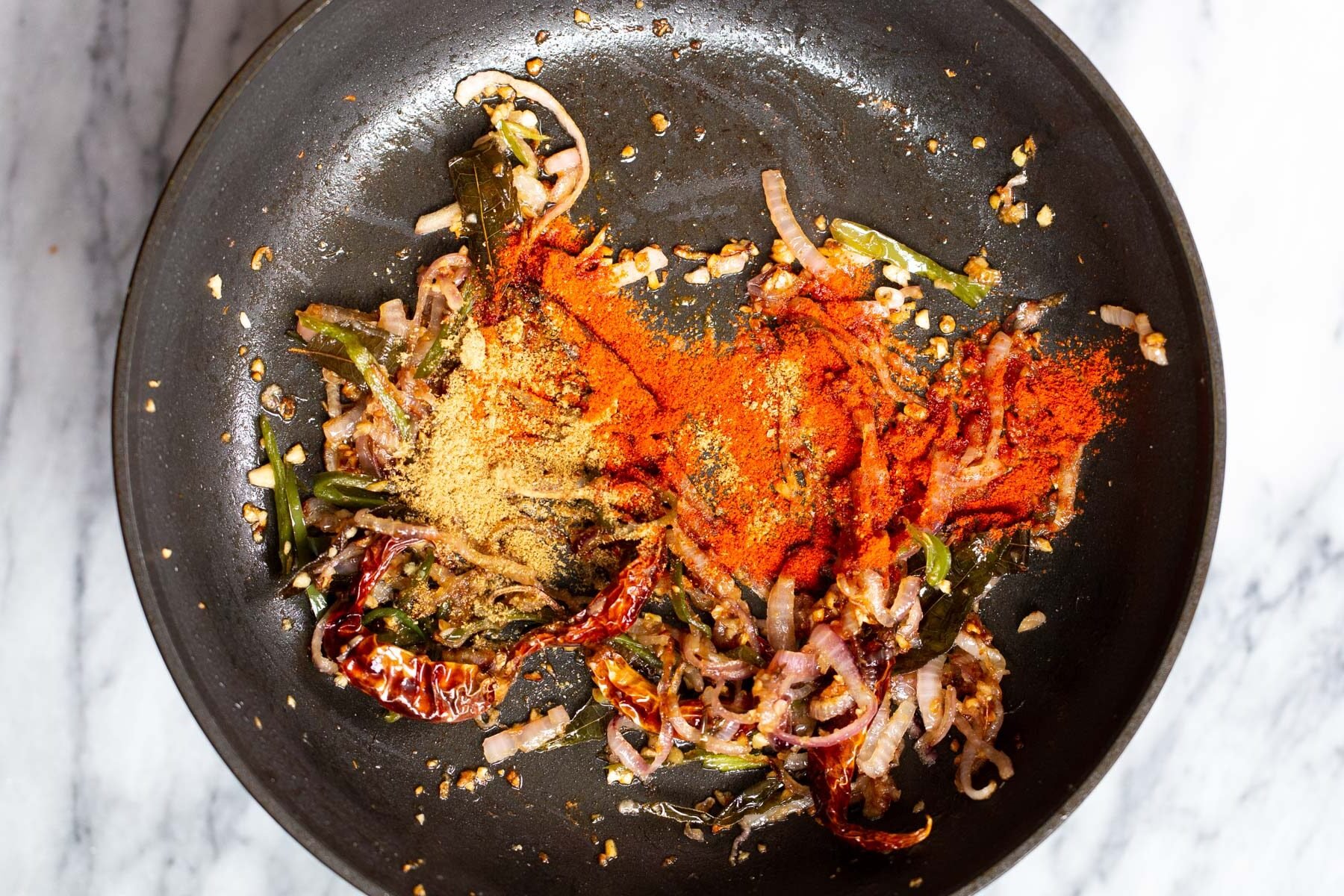chili powder being added to a frying pan sauteed garlic and onions