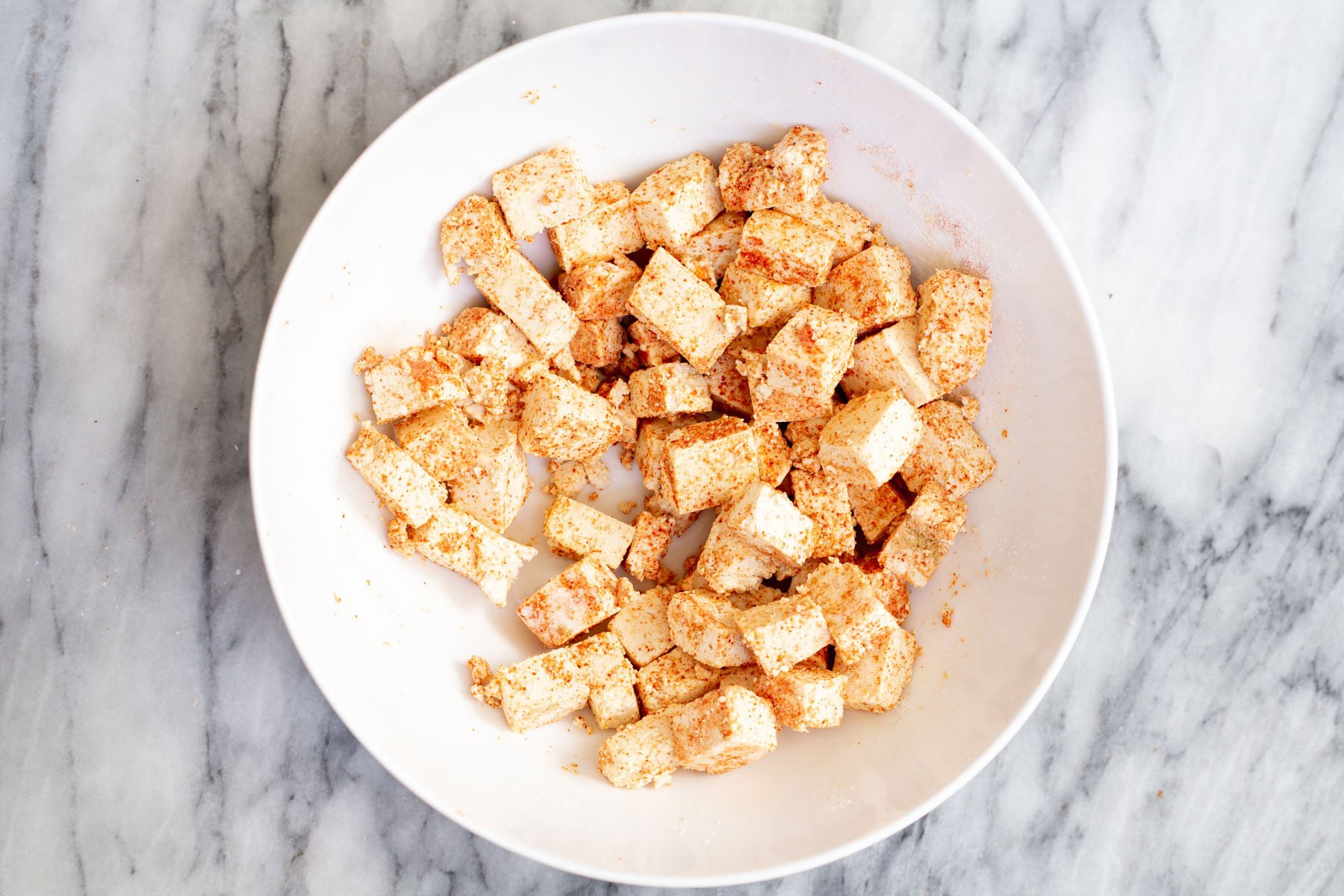 tofu cubes being tossed with cornstarch spice mix in a white bowl 