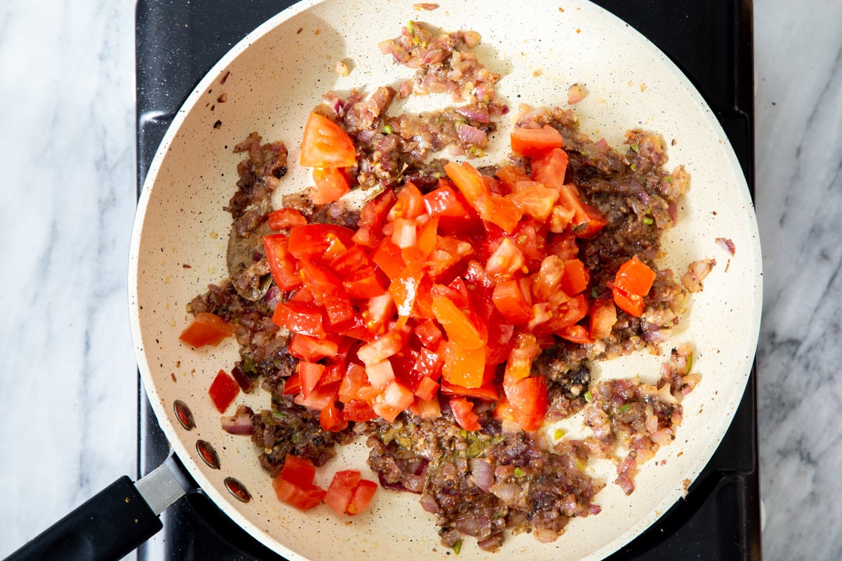 tomatoes being added to a frying pan with spices and sauteed onions for making gravy