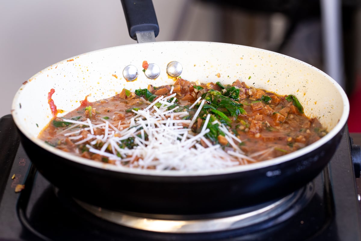 grated vegan parmesan being added to a sauteeing pan with spinach and tomato