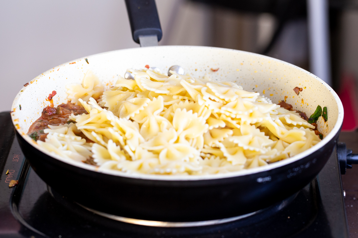 cooked vegan farfalle being added to a pan with sauteed pepperoni pasta sauce