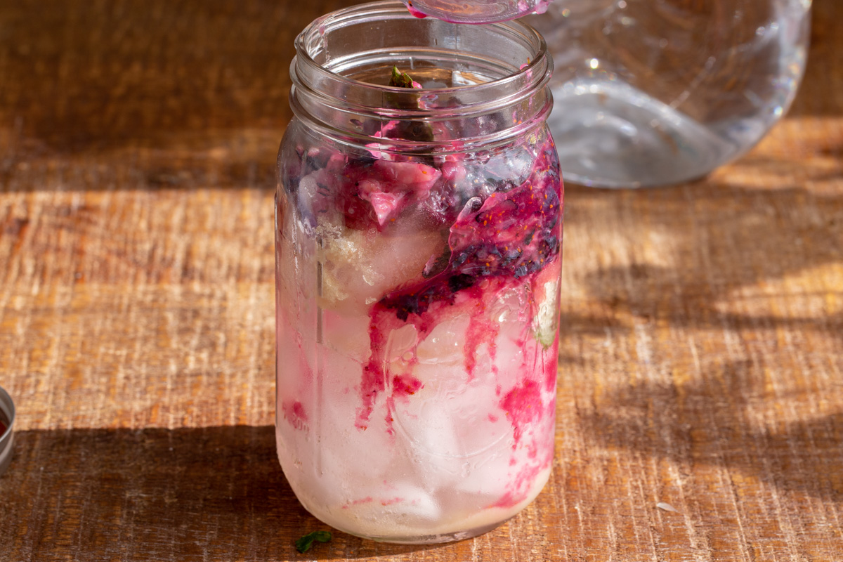 muddled blueberries being added to a mason jar with ice cubes and lemon juice to make lemonade