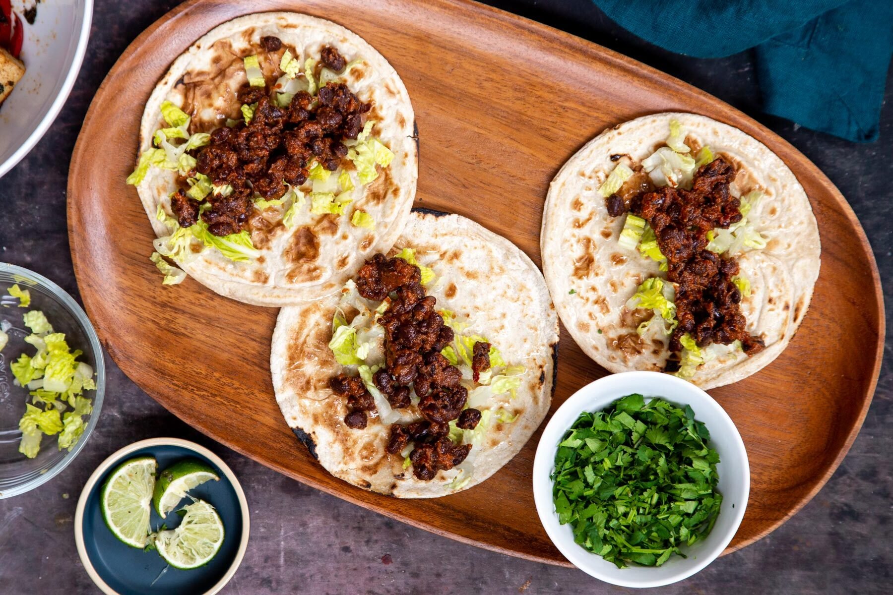 three small wheat tortillas on a wooden platter being topped with lettuce and black beans