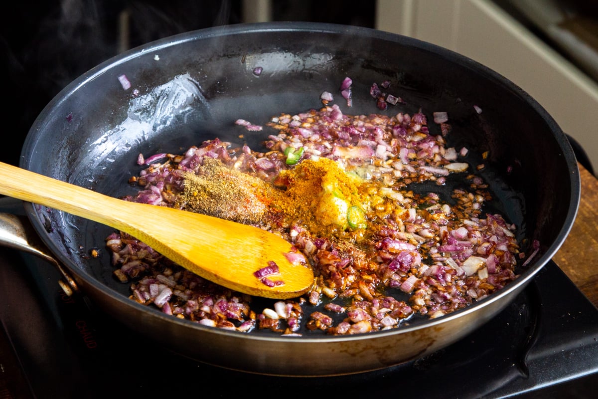 Indian spices being added to a frying pan with sauteed onions
