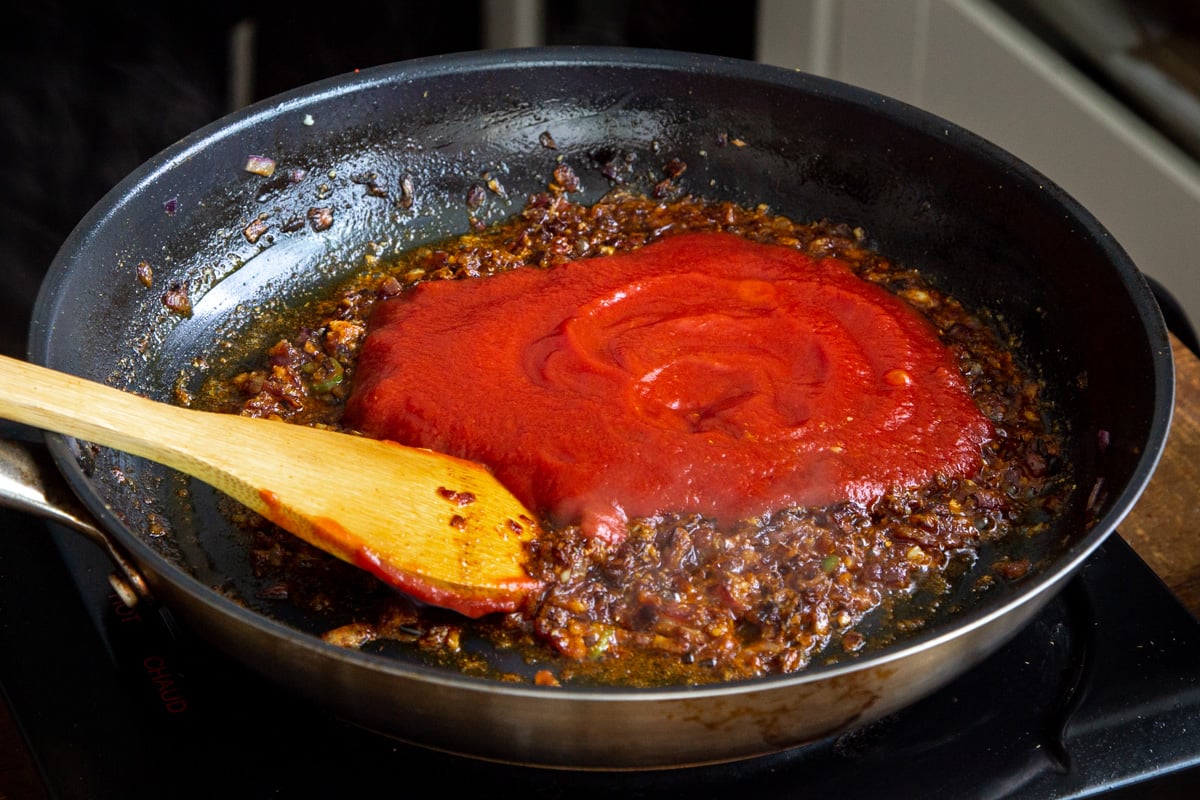 tomato puree being added to spiced onions in frying pan