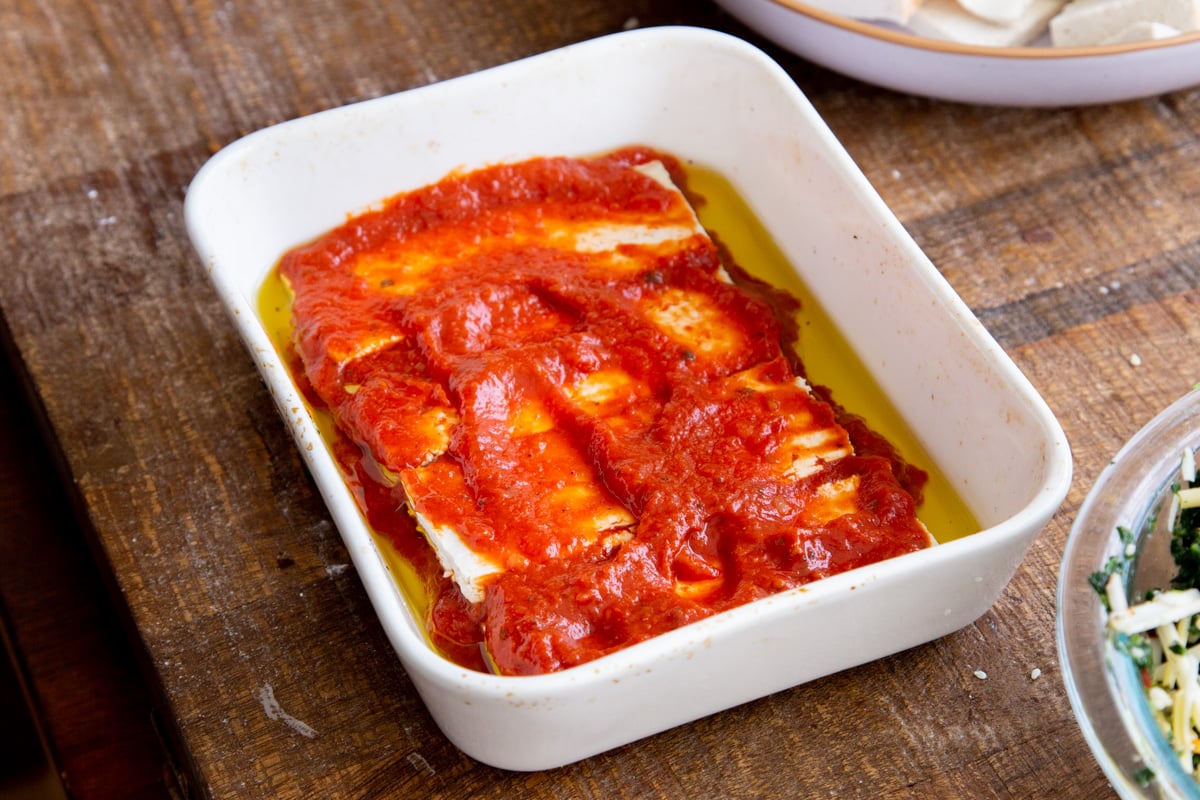 slabs of pressed tofu layered with marinara sauce and olive oil in a casserole dish