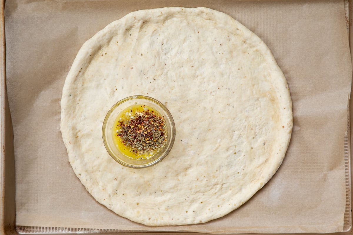 shaped unbaked vegan pizza crust with a small dish of seasoned oil on a baking sheet