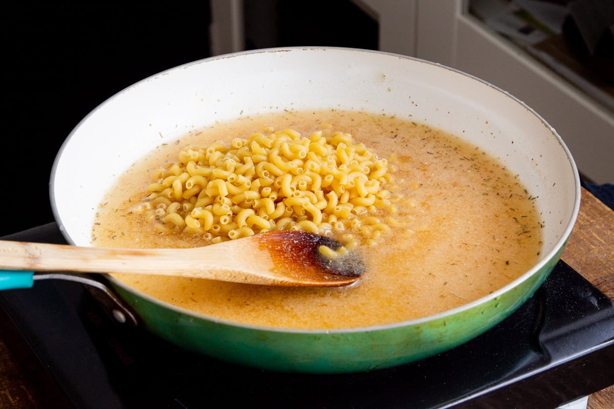elbow pasta being added to a skillet with seasoned broth
