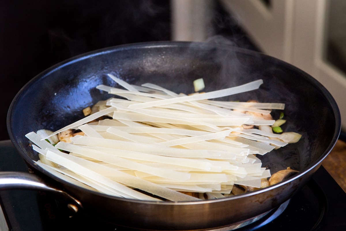 Thai rice noodles being added to a black skillet with mushrooms chili and garlic