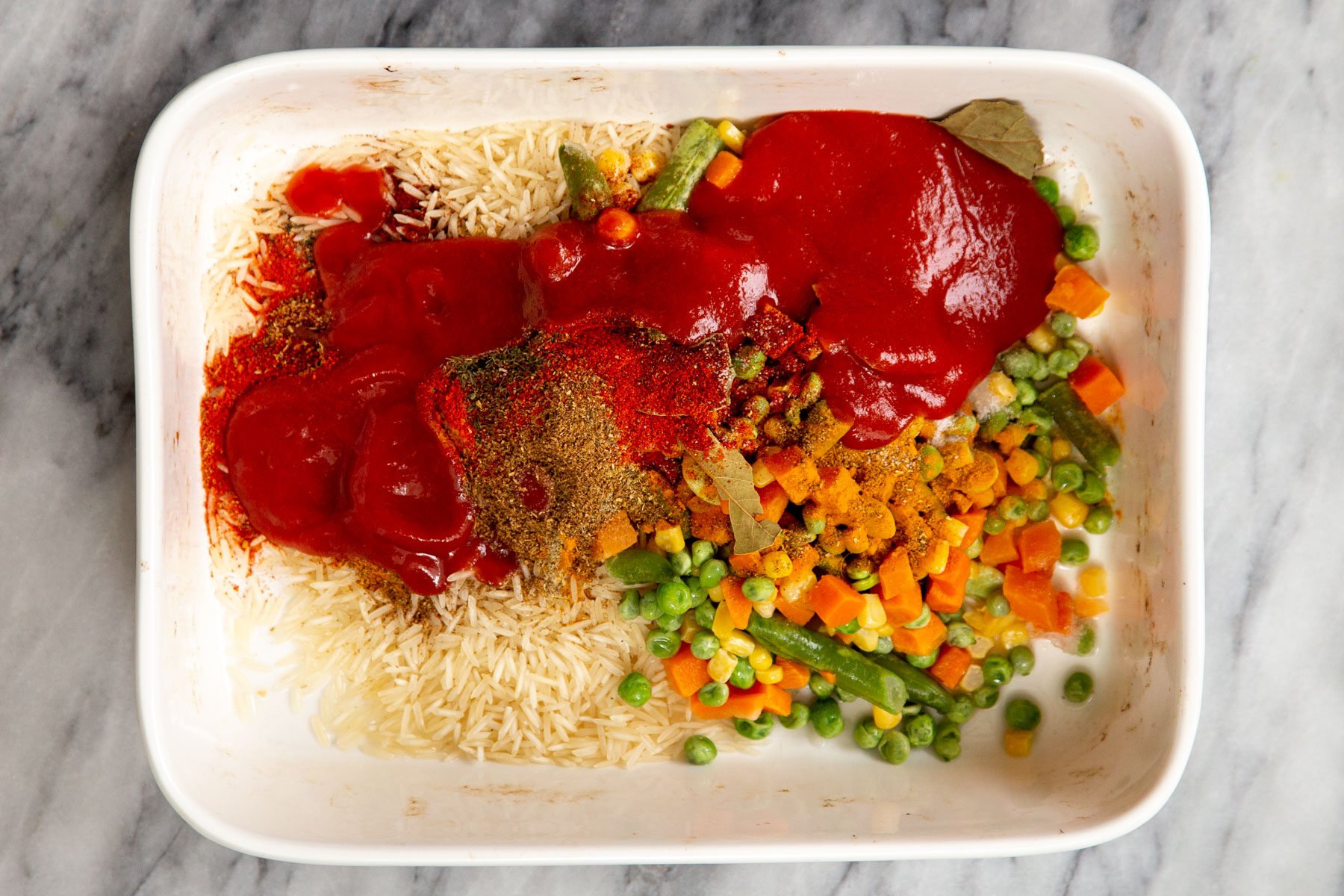 a white casserole dish with rice, tomato paste, veggies and spices