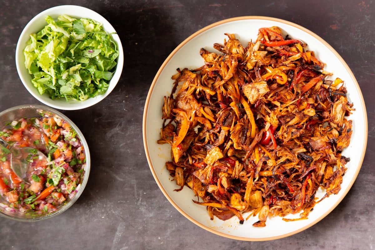 baked jackfruit taco meat in a white bowl next to two small bowls with chopped lettuce and salsa