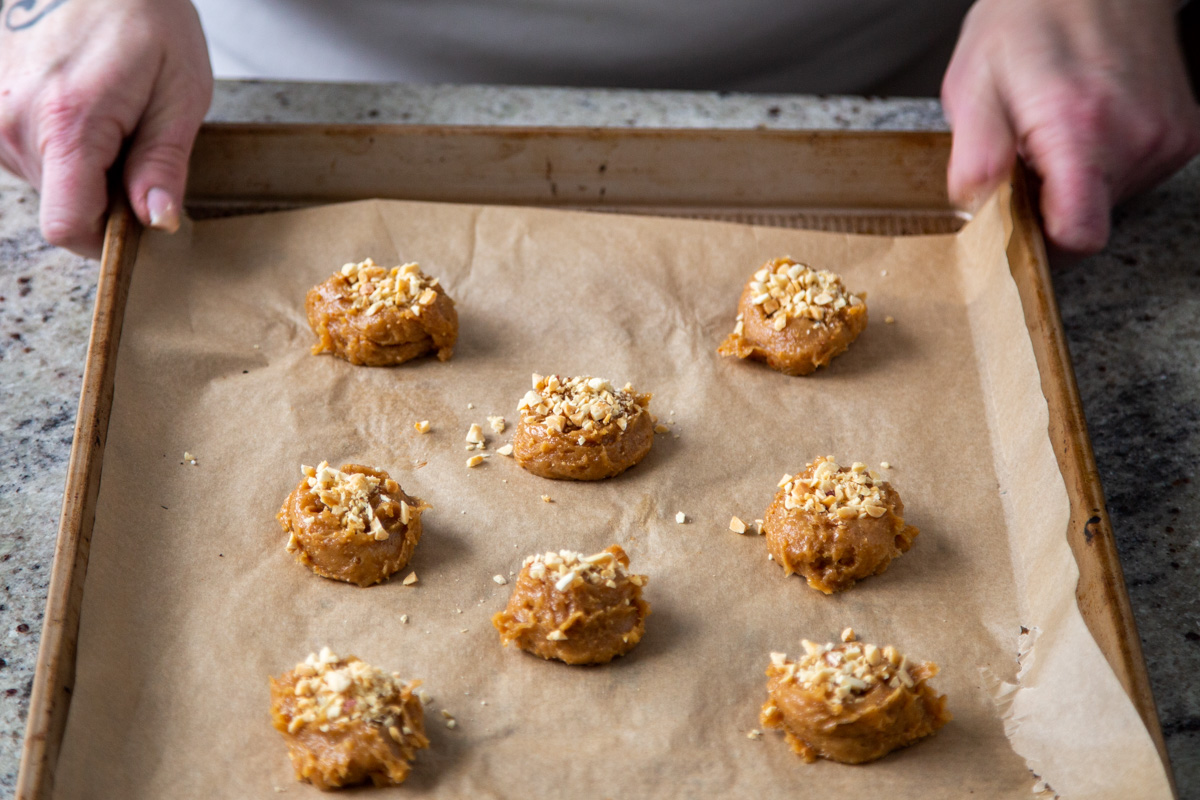 crushed peanuts being sprinkled on peanut butter cookie batter on a baking sheet