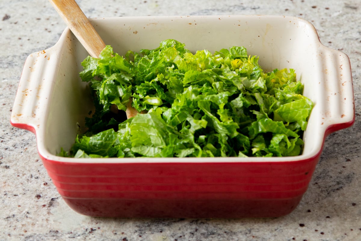chopped mustard greens being added to a red casserole dish