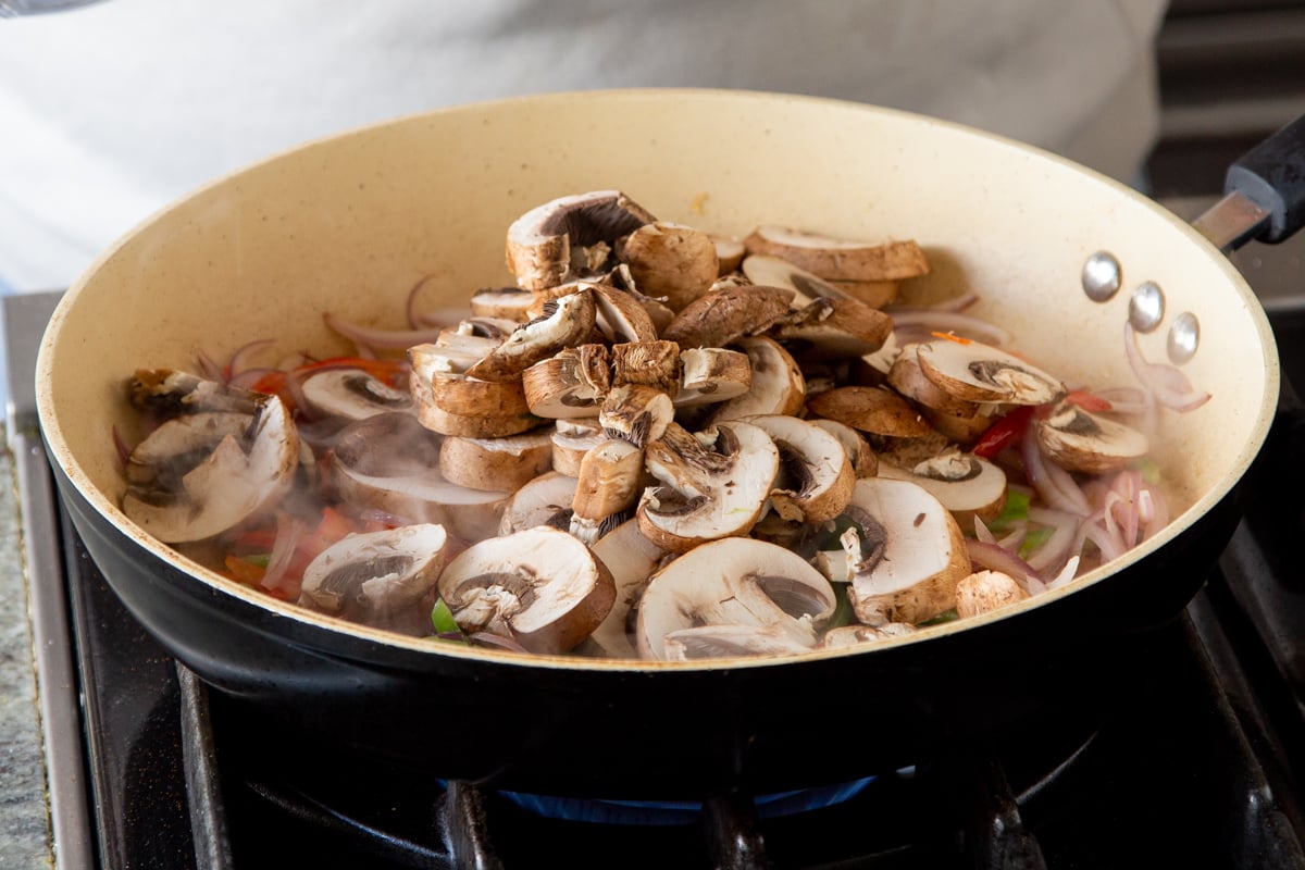 sliced mushrooms being added to a sauteeing pan with sliced bell peppers and onions