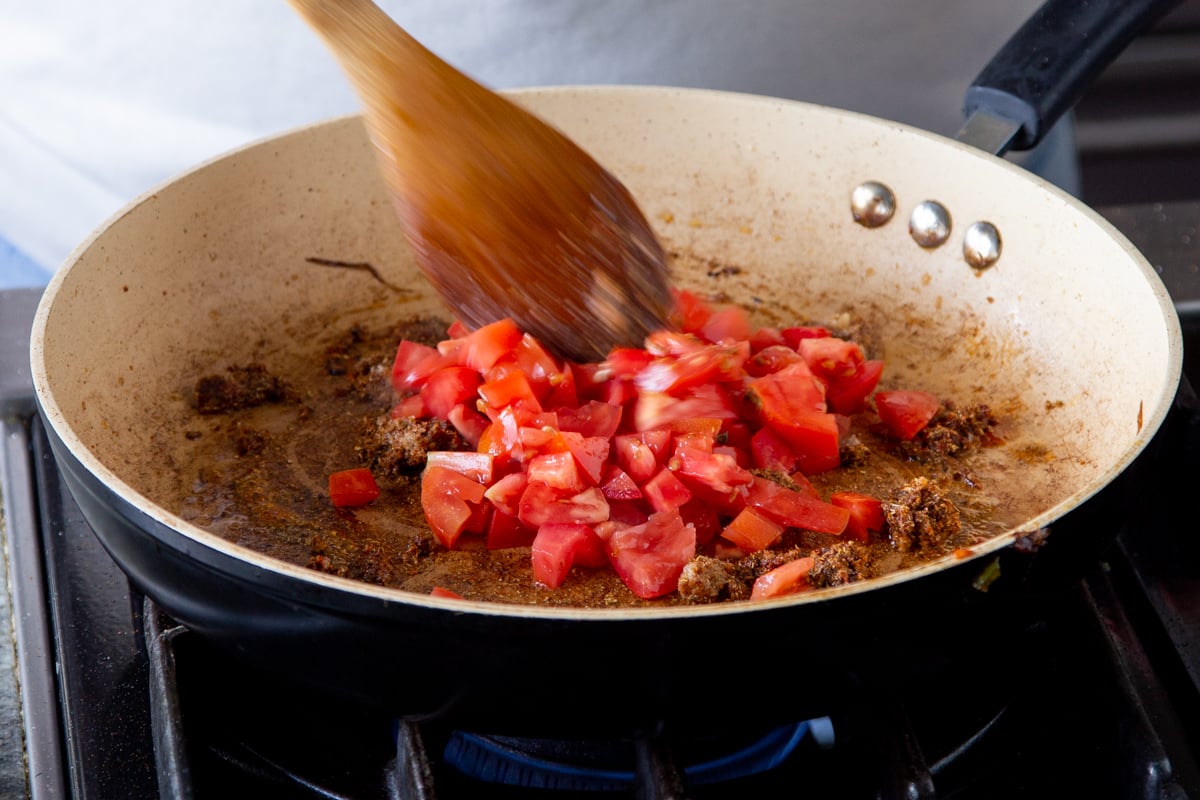 chopped fresh tomatoes being added to a sauteeing pan with spices