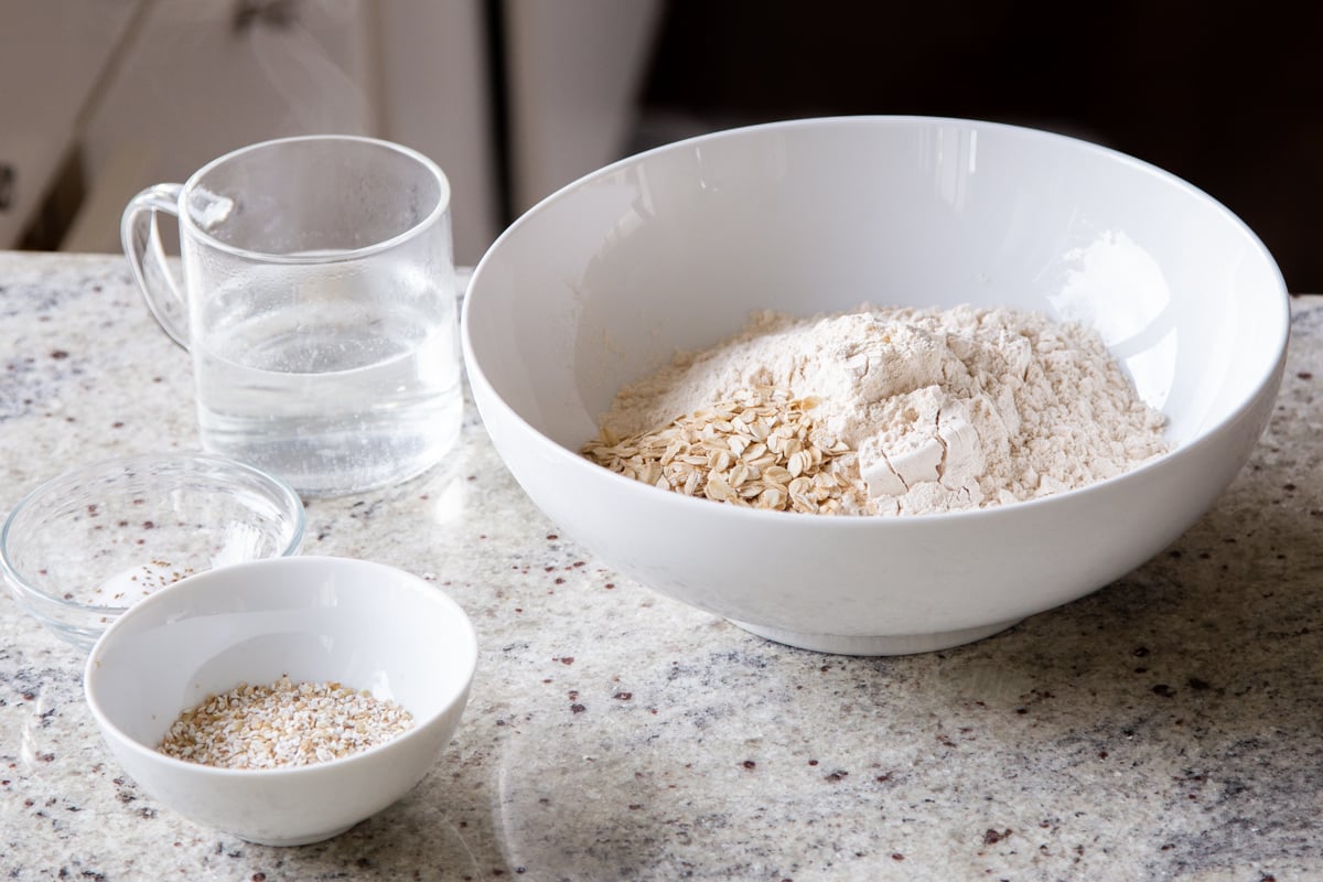 flour and whole grains in a white mixing bowl next to a bowl of buckwheat and a cup of warm water