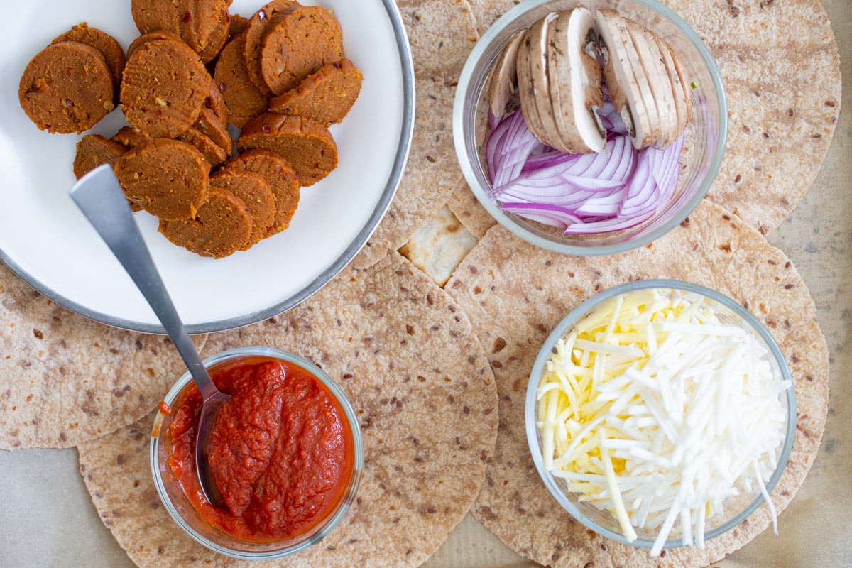 vegan pepperoni, vegan cheese, pizza sauce, and veggies in bowls arranged on top of tortillas