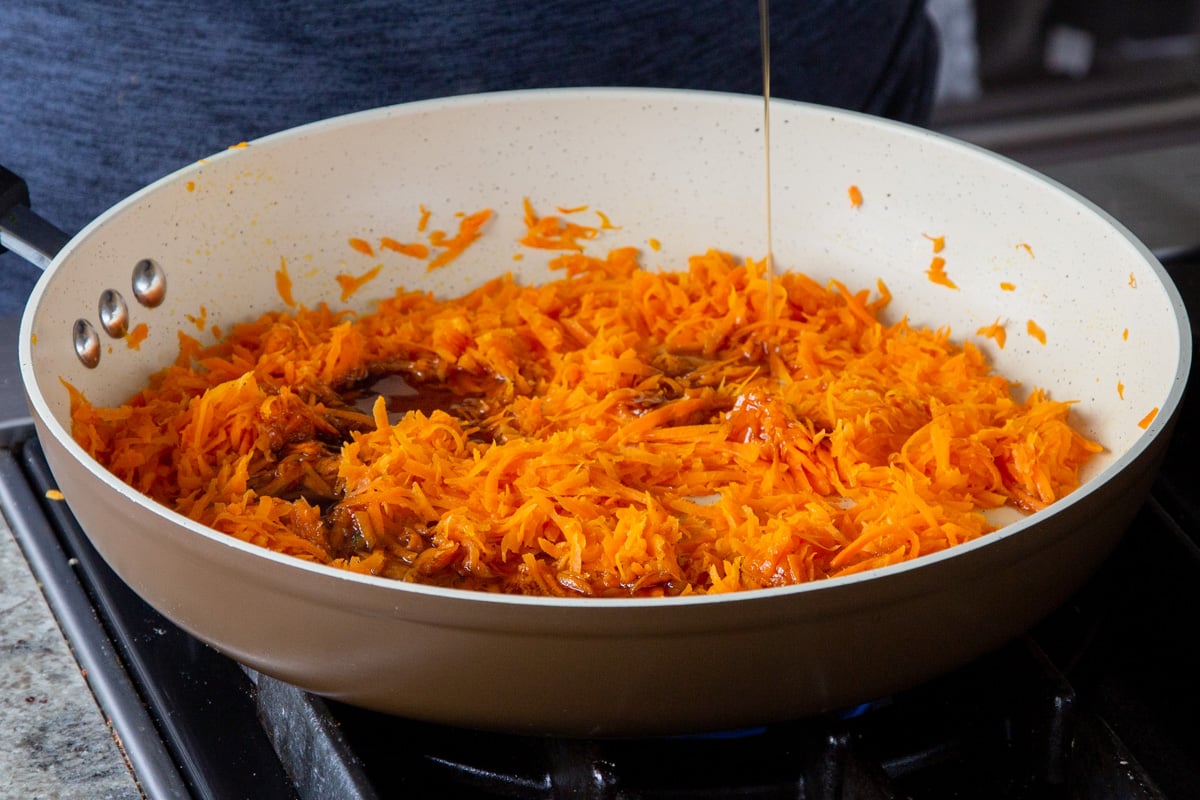 adding maple syrup to the pan of shredded carrots