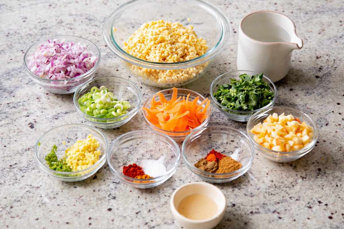 soaked moong dal with fruit, veggies, and seasonings in bowls on a kitchen counter