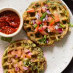 two spinach waffles on a plate topped with chopped tomato and cilantro with red dipping sauce next to them