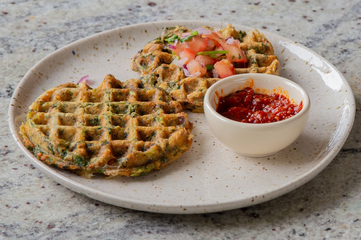 two spinach waffles on a plate topped with chopped tomato and cilantro with red dipping sauce next to them