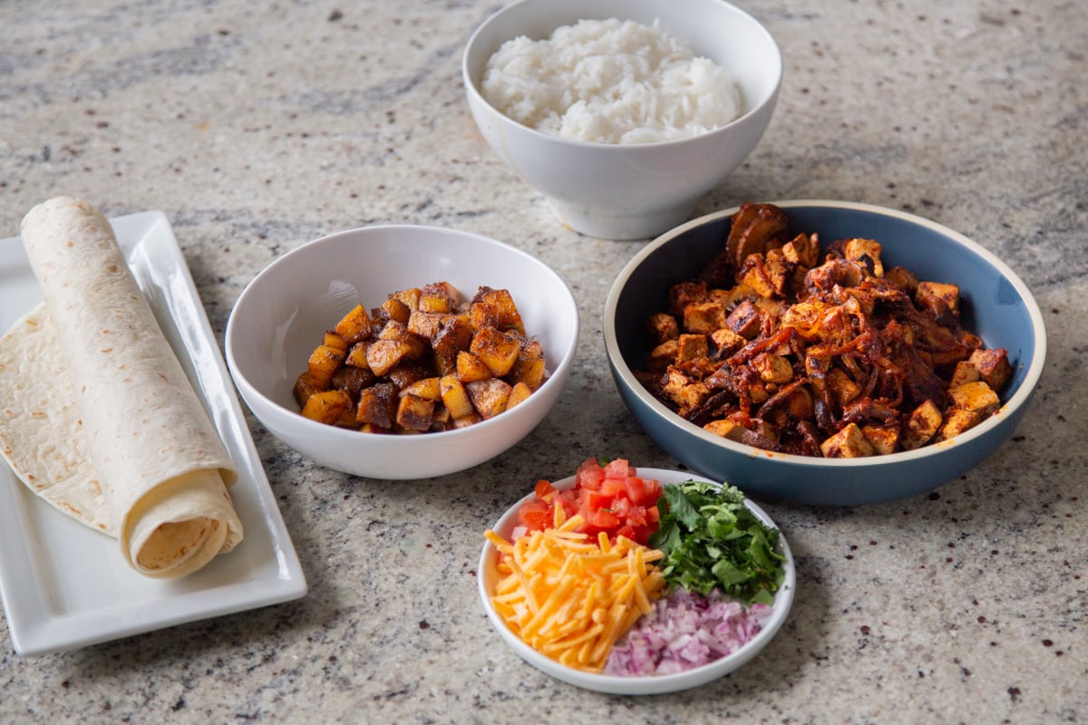 rice, spicy potatoes, tofu mushrooms mixture, toppings, and tortilla in bowls on a kitchen counter